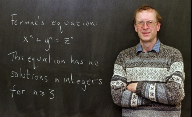 Andrew Wiles, a mathematician with glasses, standing in front of a chalkboard displaying Fermat's equation. He is wearing a striped shirt and patterned sweater, smiling gently.