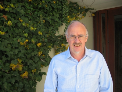 A headshot of David Masser, a mathematician, smiling gently. He has light-colored hair, glasses, and a short beard, with a green ivy-covered wall in the background.