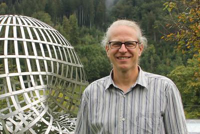 Jakob Stix, a mathematician, smiling outdoors with a metallic sculpture in the background. He has light-colored hair, dark-rimmed glasses, and is wearing a striped collared shirt.
